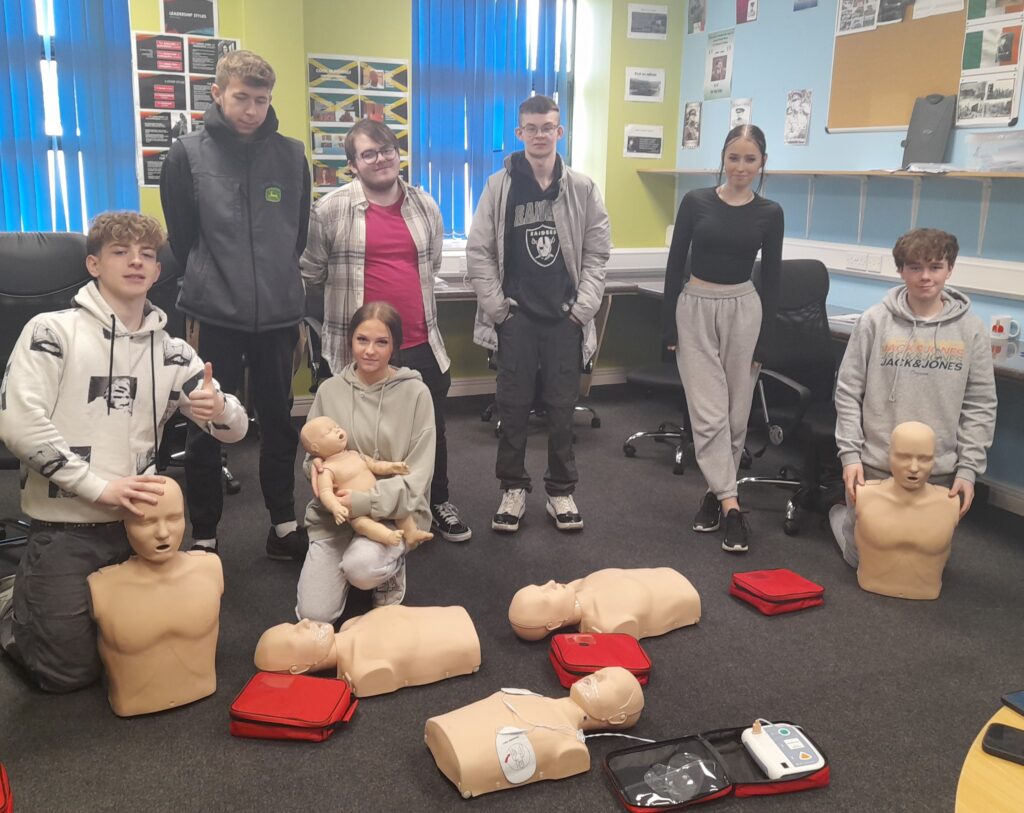 Youthreach students in a colourful classroom setting. Three students are kneeling on the floor holding first aid dummies and four students standing behind.