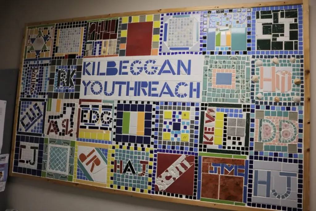A vibrant mosaic sign on the wall reads KILBEGGAN YOUTHREACH in bold blue letters, framed by an array of colourful, patterned tiles arranged in striking geometric designs.