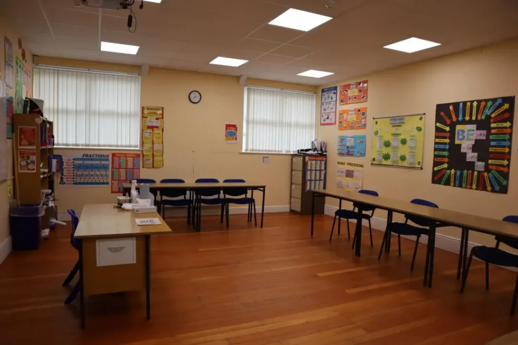 A classroom on the Ballymahon campus with wooden floors, several rectangular tables and blue chairs, educational posters on the walls, and windows with blinds letting in natural light. A small clock is mounted on the wall.