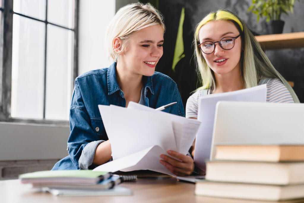 Two young women studying together at a desk, using learner support resources as they hold papers and look at a laptop screen, with books and notebooks on the table in a bright room.