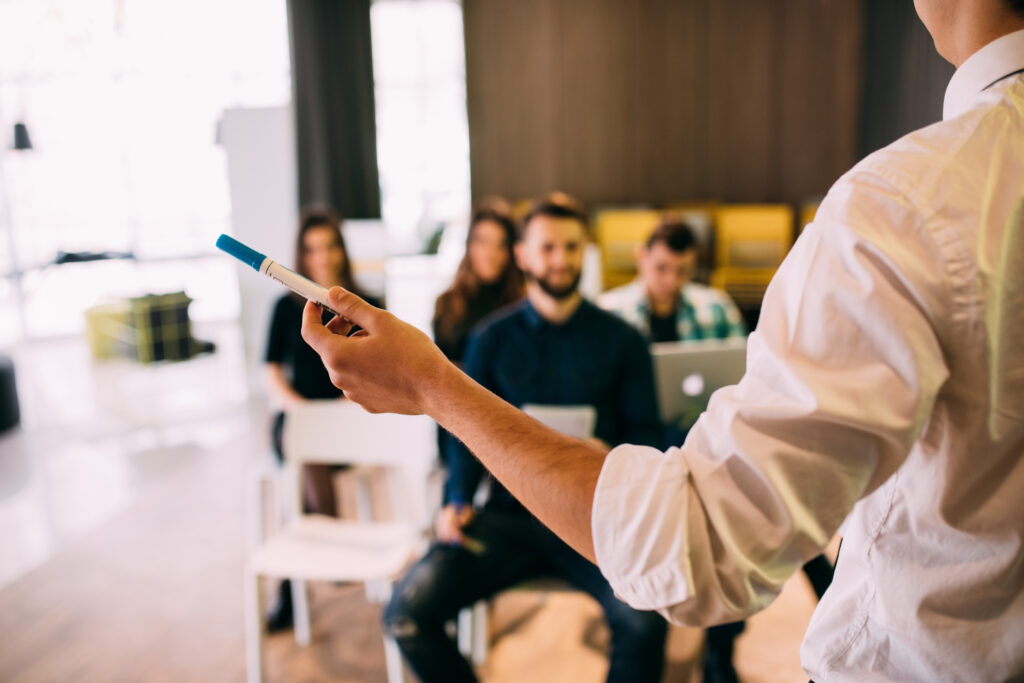 A person in a white shirt holding a marker is presenting Skills to Advance to a group of seated people, some with laptops, in a bright, modern room. The focus is on the presenter’s hand and marker, with the audience blurred in the background.
