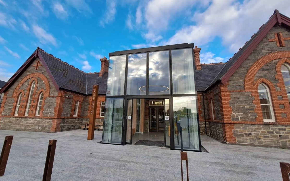 Modern glass entrance with a circular light hangs between two sections of a traditional stone and brick building, under a blue sky with scattered clouds.