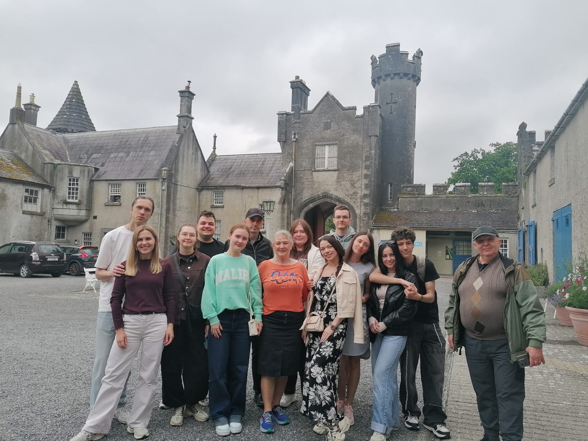 A group of thirteen people pose together in front of a stone building with a tower and arched entrance on a cloudy day.